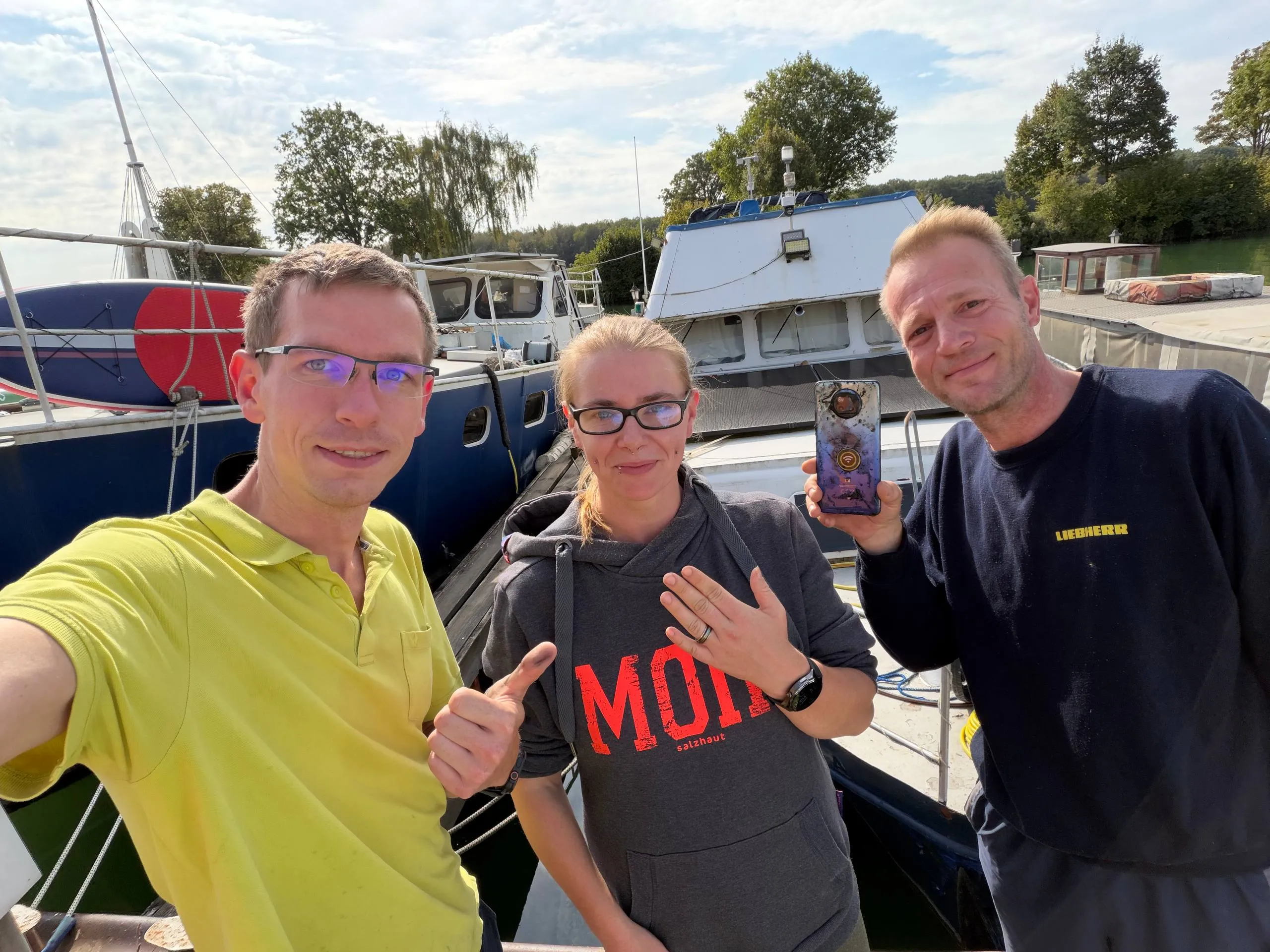 Drei Personen auf einem Steg am Hausboothafen in Hannover, in der Mitte eine Frau mit Ehering am Finger, rechts ein Mann, der ein geborgenes Handy in die Kamera hält.