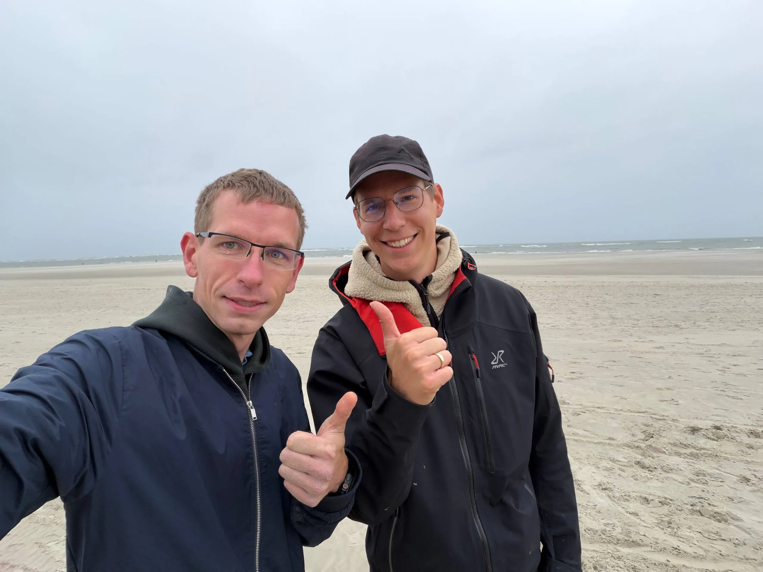 Zwei Männer am Nordseestrand von Spiekeroog zeigen lächelnd den Daumen nach oben, im Hintergrund Watt und Meer.